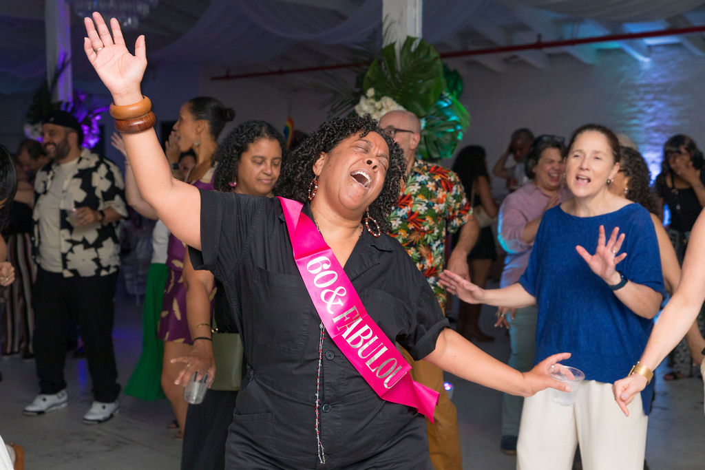 The 60-year-old honoree — arms wide open, sash blazing pink, the whole room dancing behind her