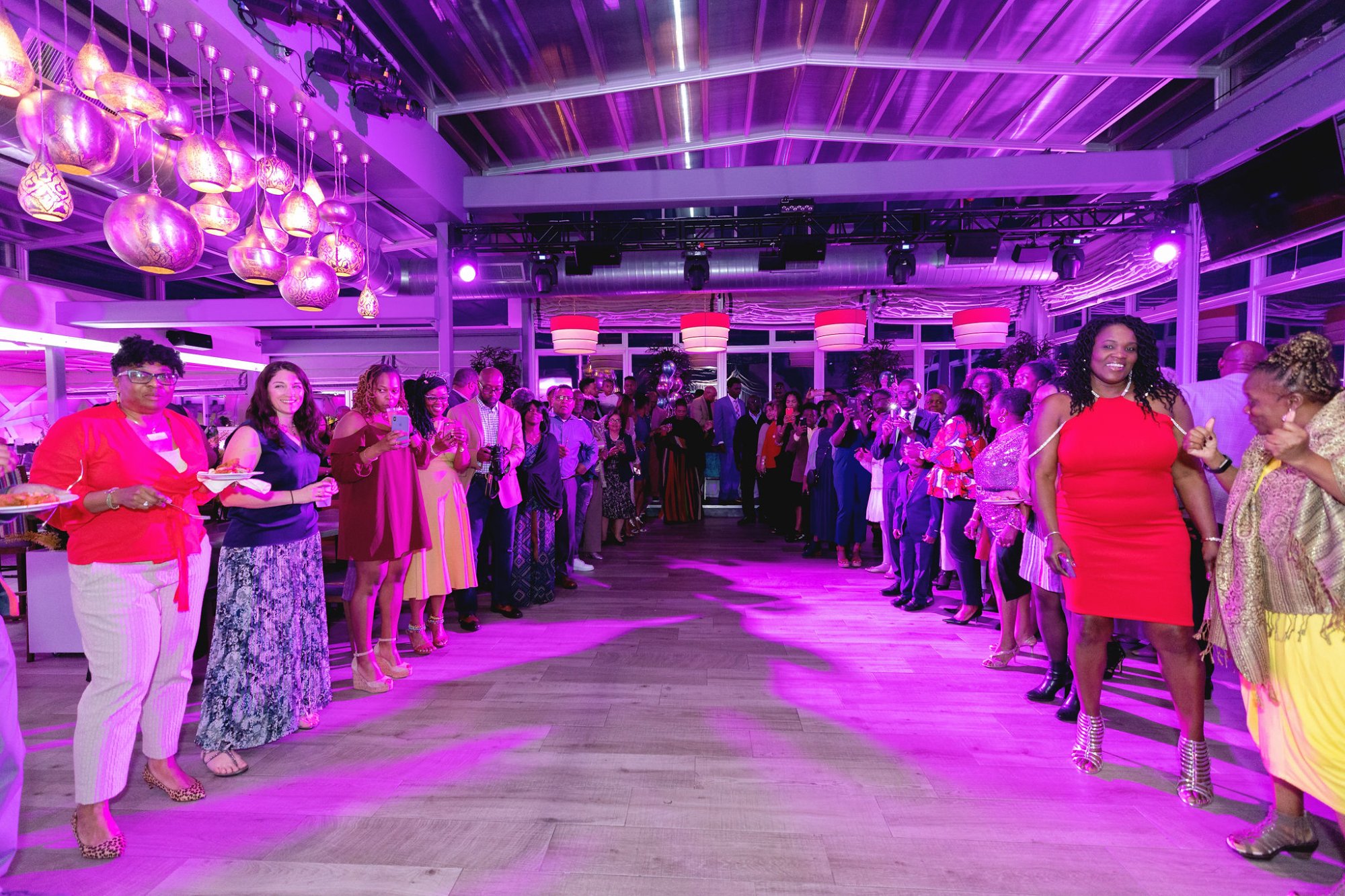 The grand entrance — a room full of guests parting to create a corridor, pink and purple light, the honoree walking in