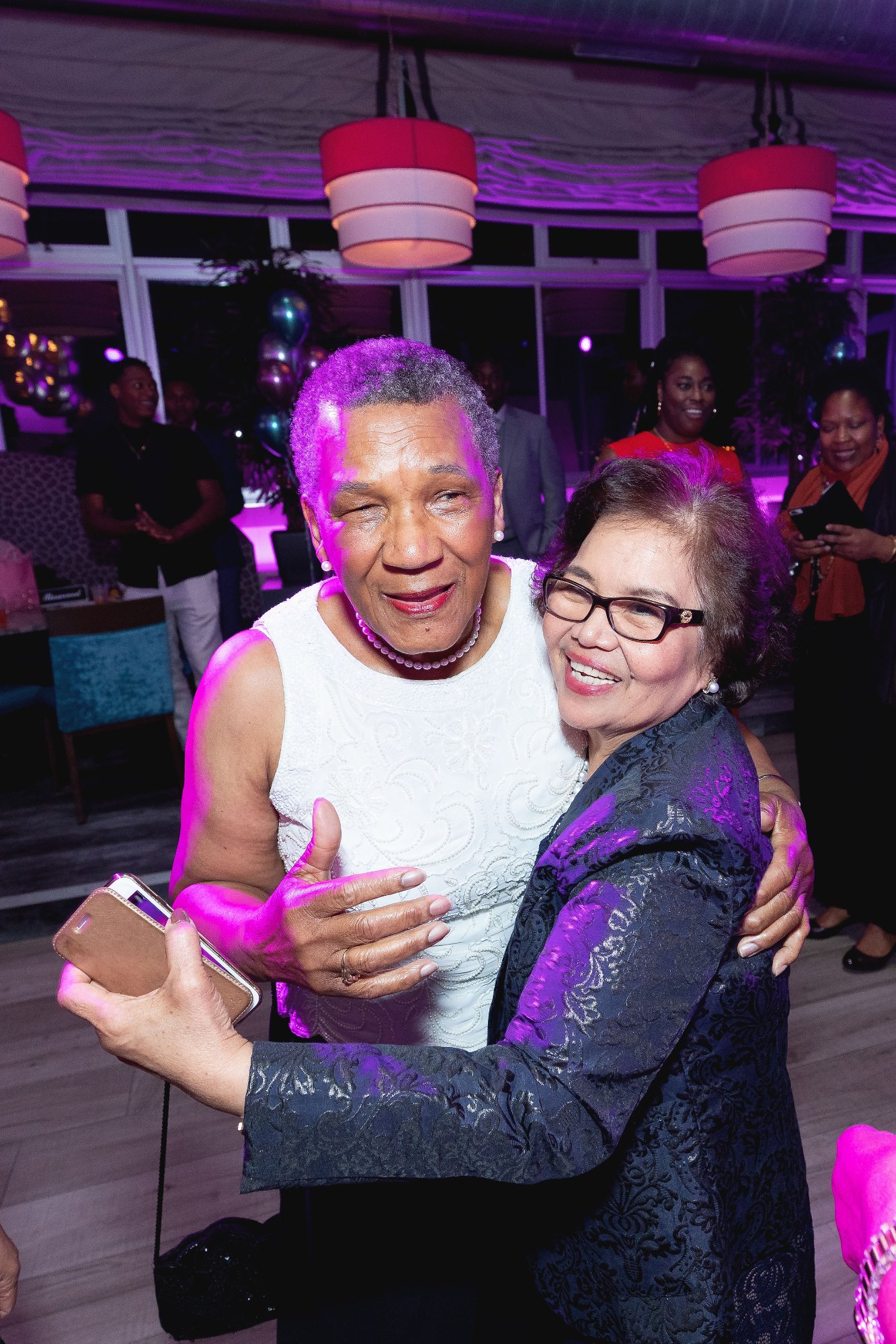 The honoree and a guest — faces close, arms fully wrapped, an embrace that holds a whole history