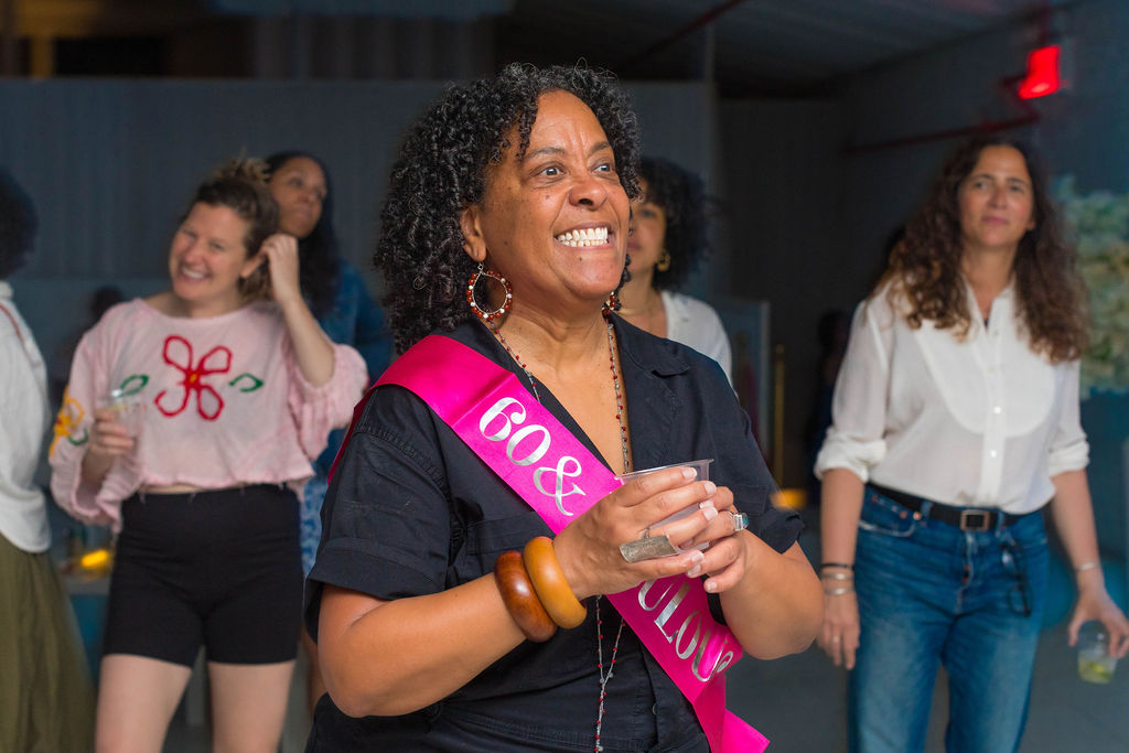 The 60-year-old honoree laughing, pink sash bright, guests behind her