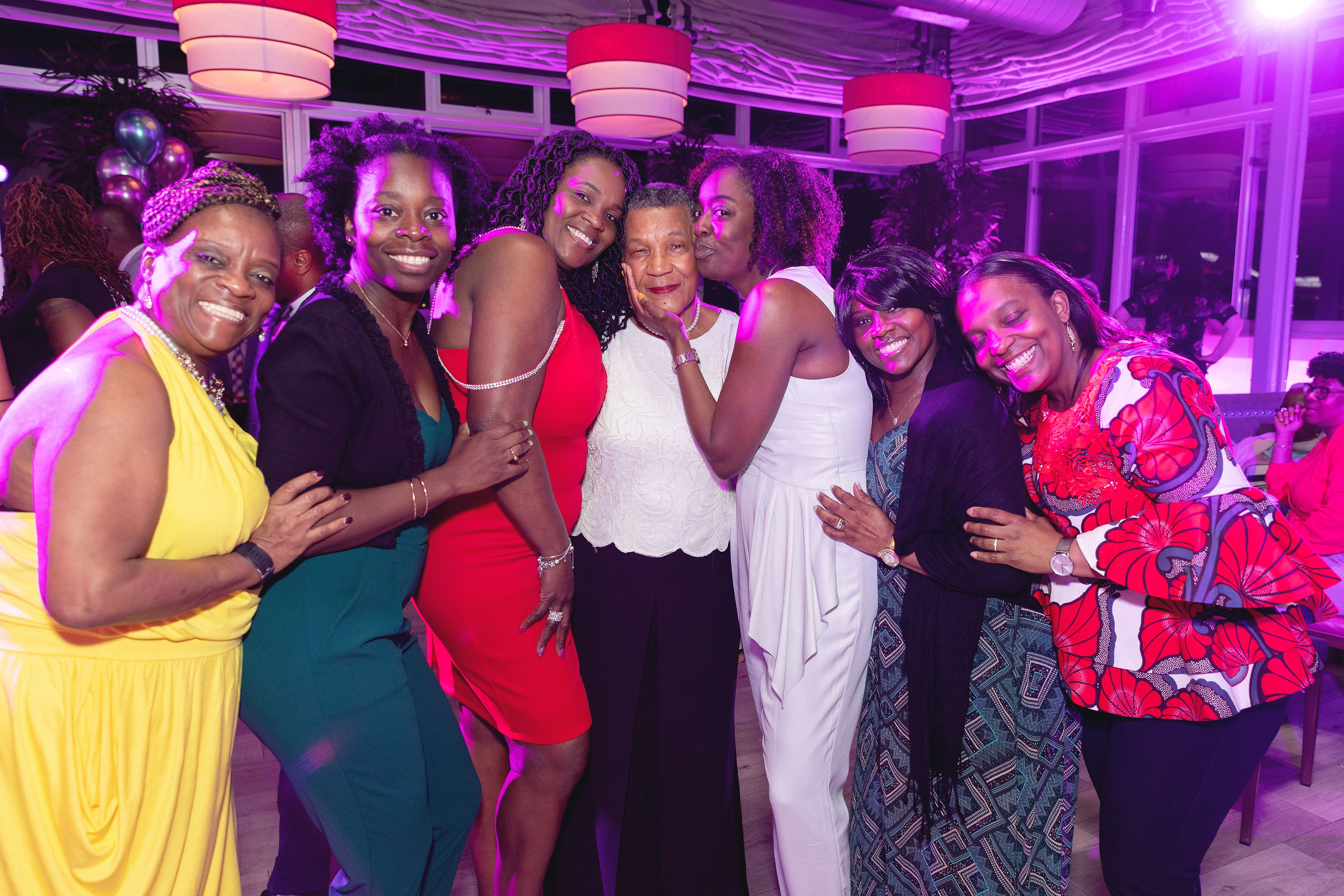 The honoree surrounded by six women — arms around each other, the room glowing behind them