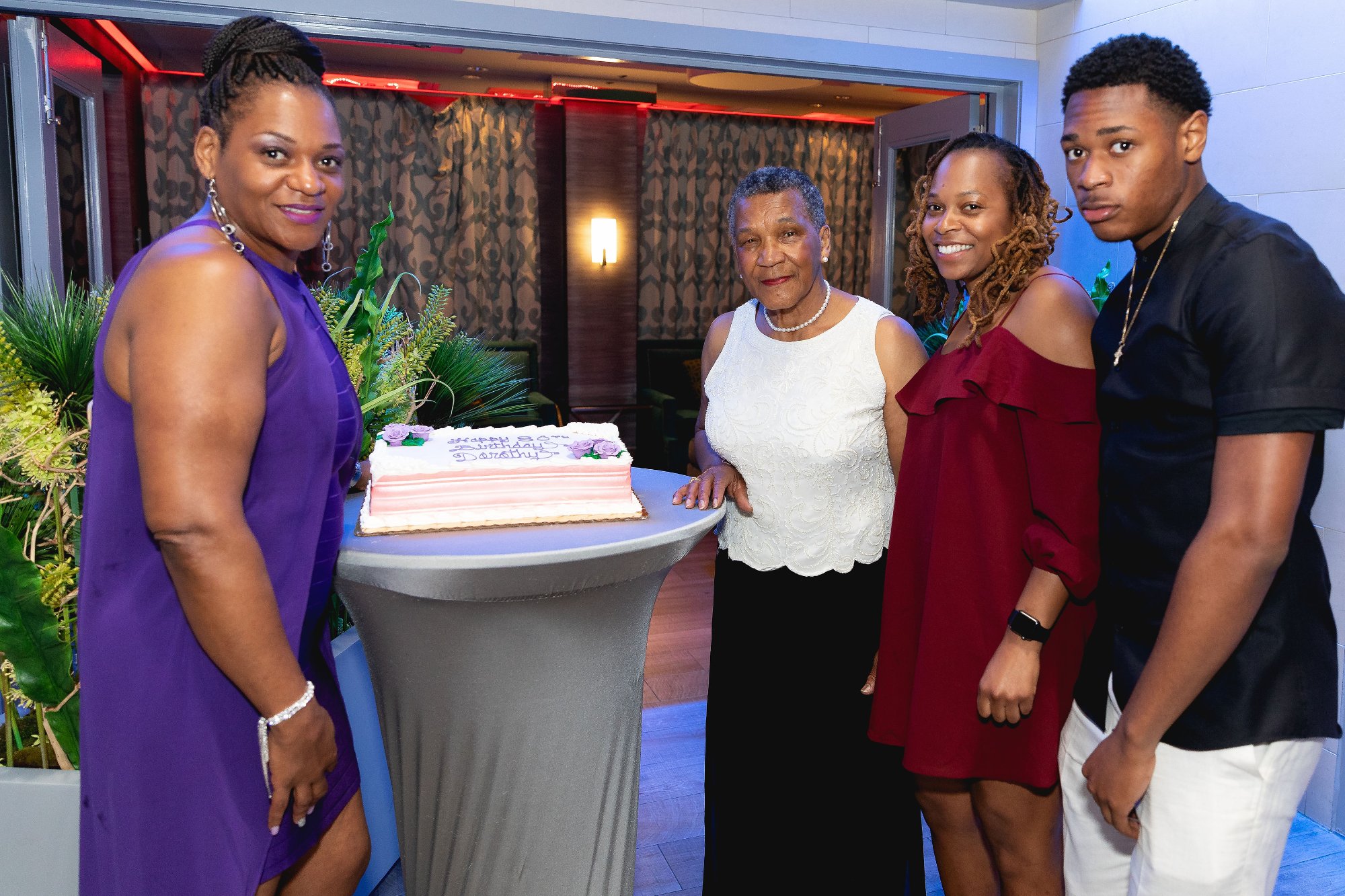 Full family photo — three generations gathered under pink light, everyone together for the honoree's 80th birthday