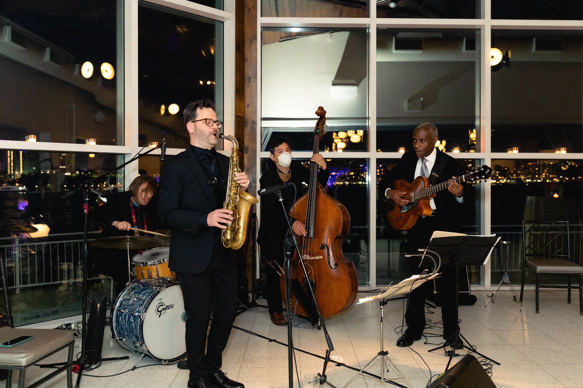 Jazz quartet performing at gala — saxophone, bass, drums, guitar — city lights behind