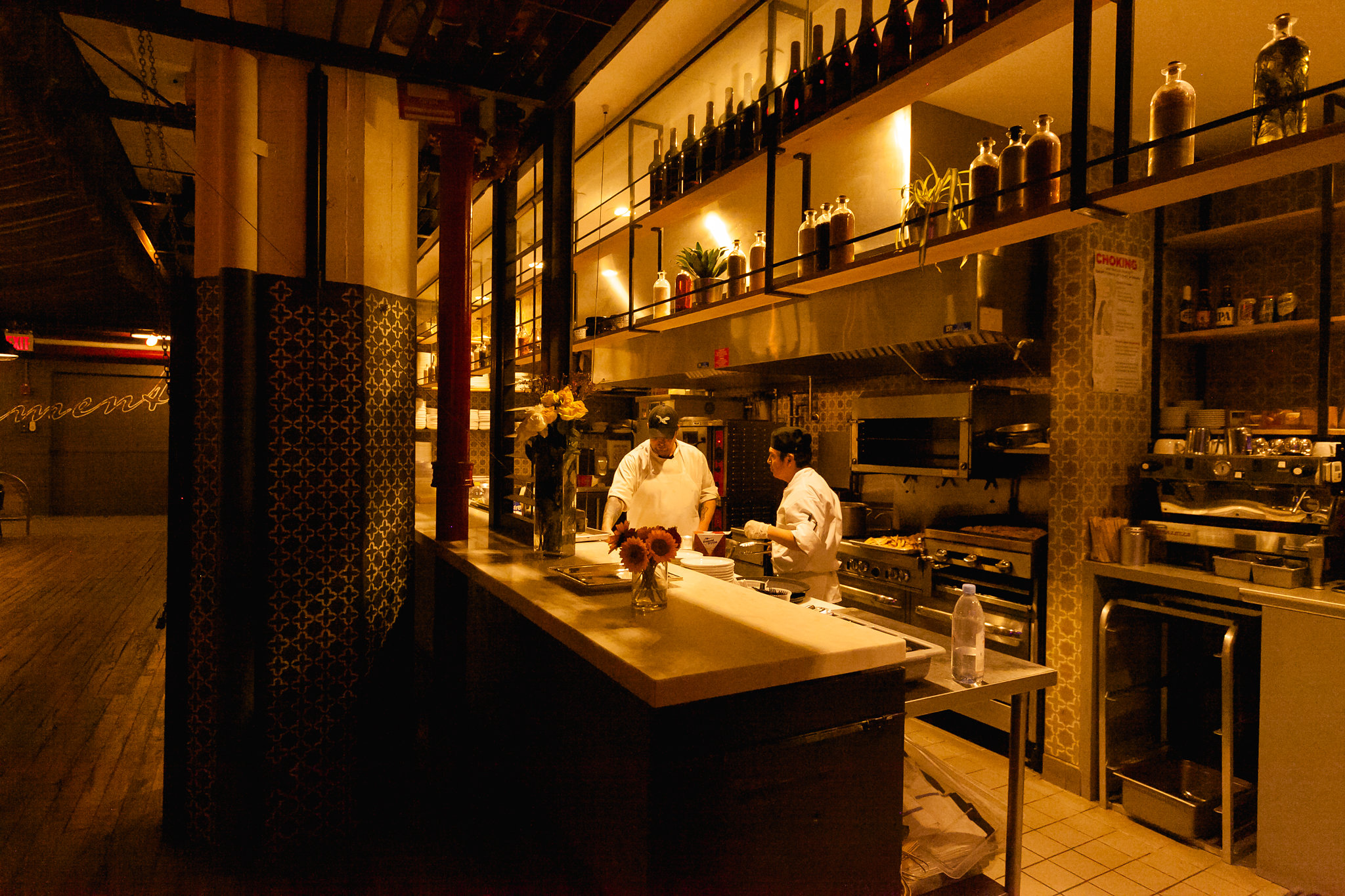 The kitchen before service — two chefs in white, warm amber light, bottles lining the upper shelves