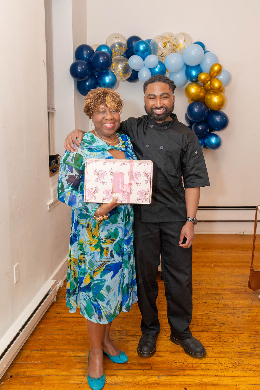 Guest and chef together holding a floral gift box — balloon arch behind them, joy between them